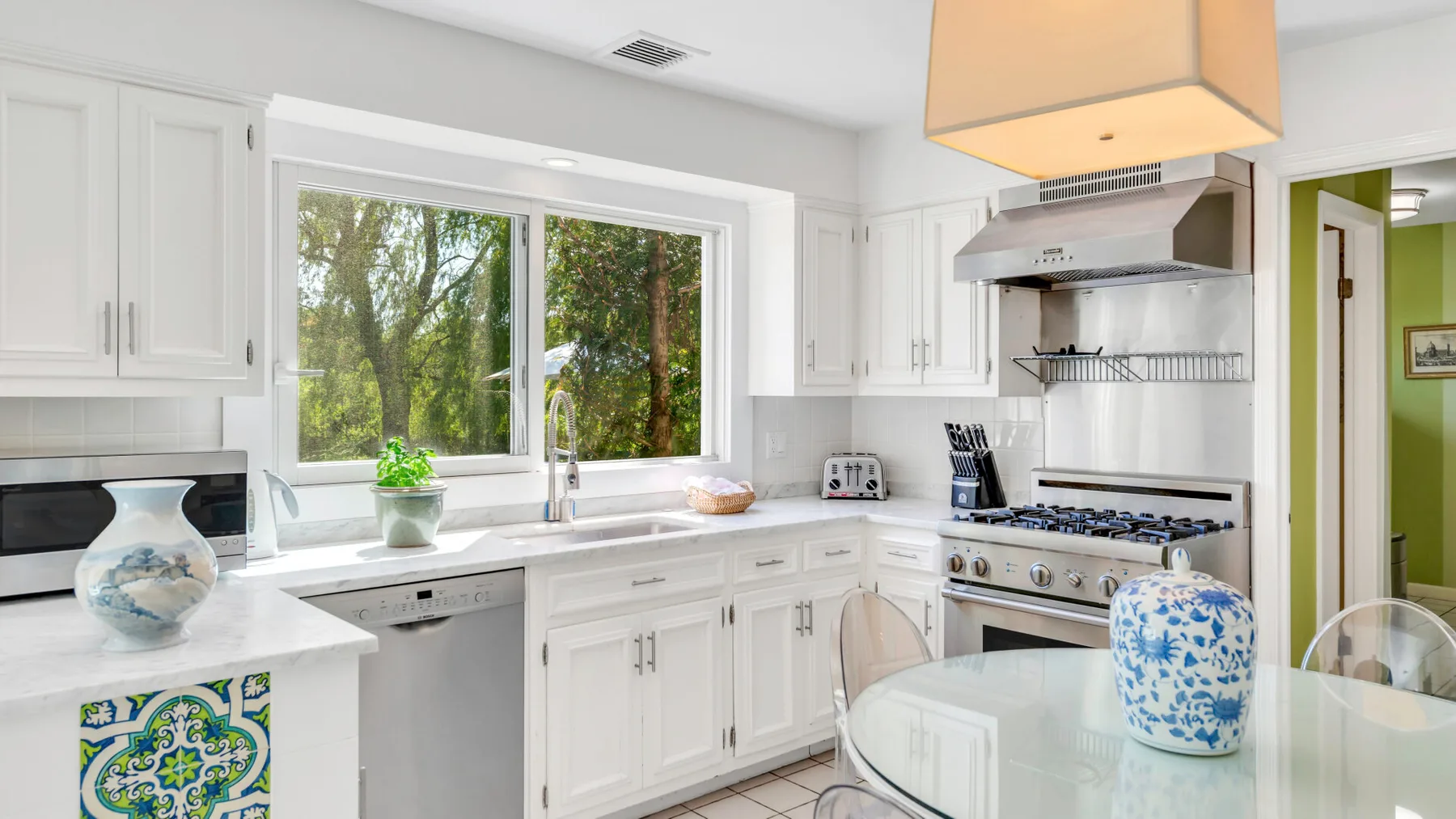 Una cocina luminosa con gabinetes blancos, una ventana grande y una mesa de comedor de vidrio.