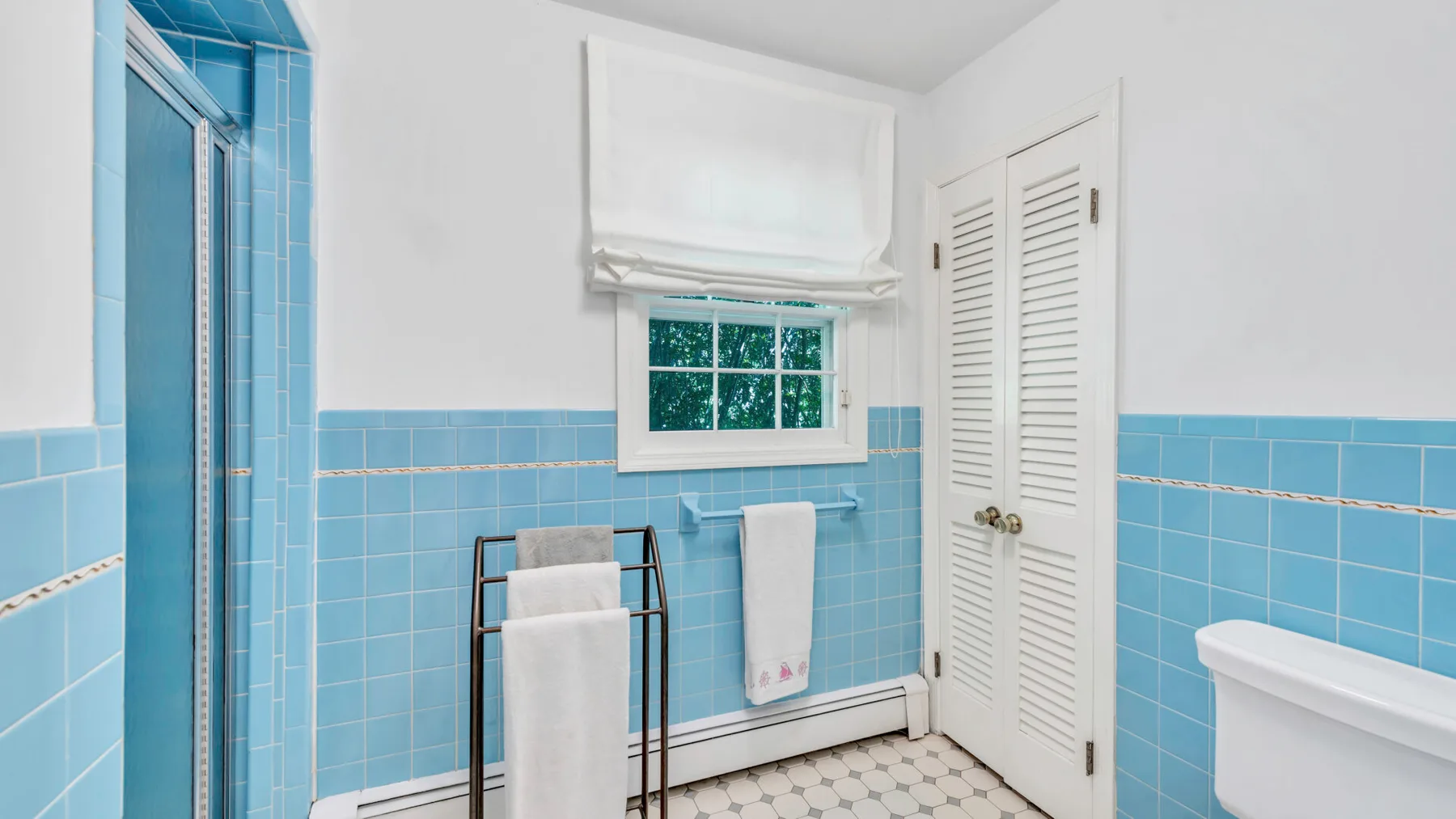 Un baño con azulejos azules, ducha, ventana y puertas blancas con celosías.