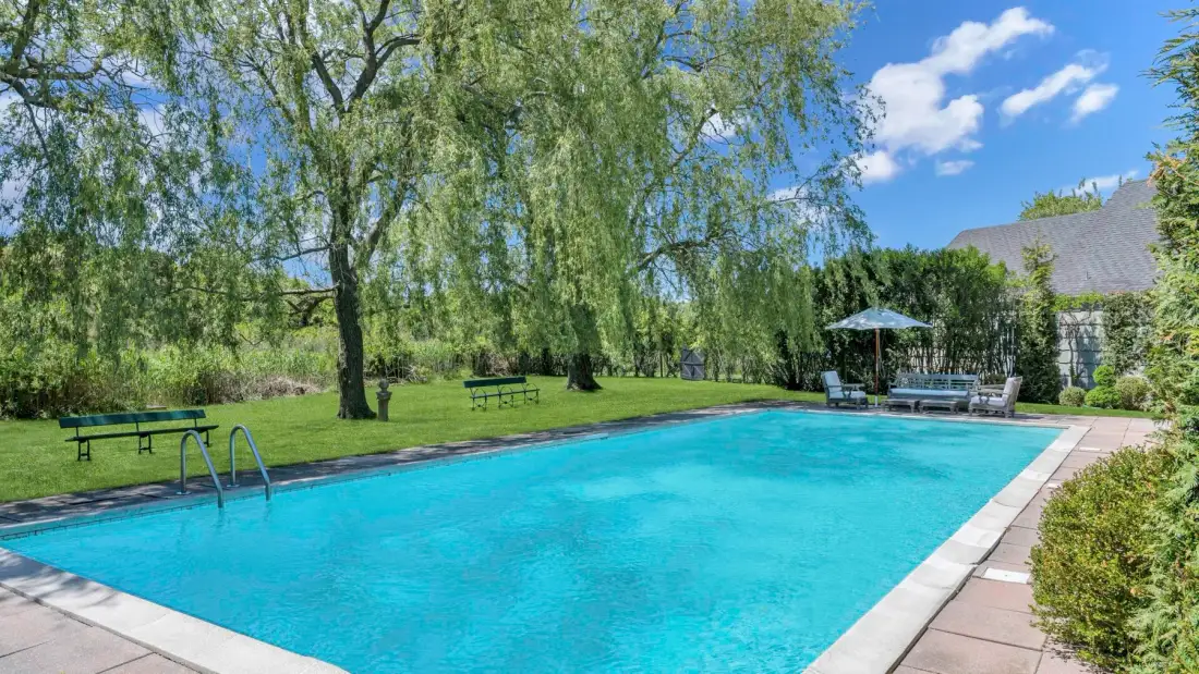An outdoor swimming pool sits beneath leafy trees, with seating along the far edge.