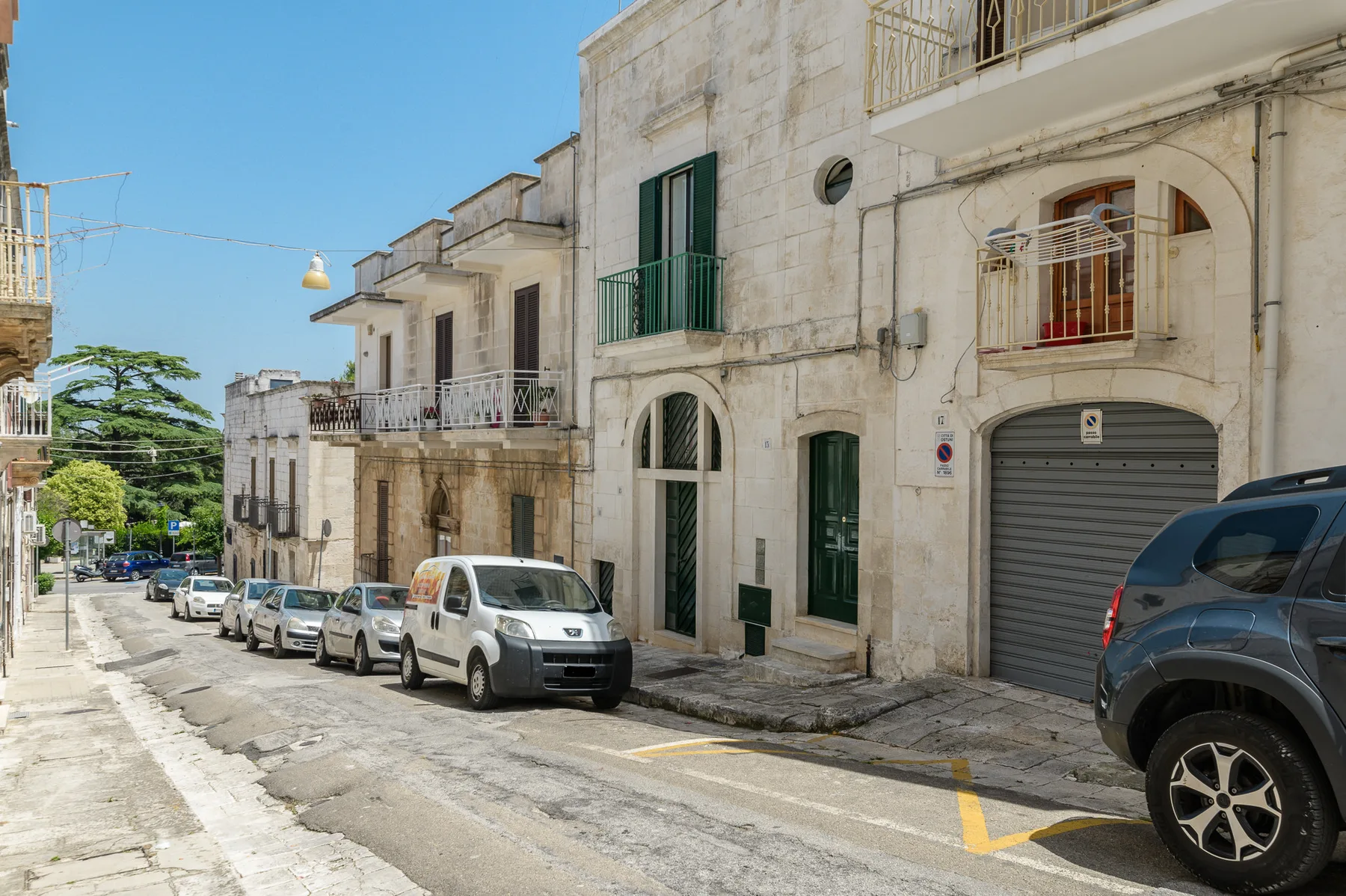 Una calle estrecha bordeada de edificios de piedra clara, balcones y coches estacionados.