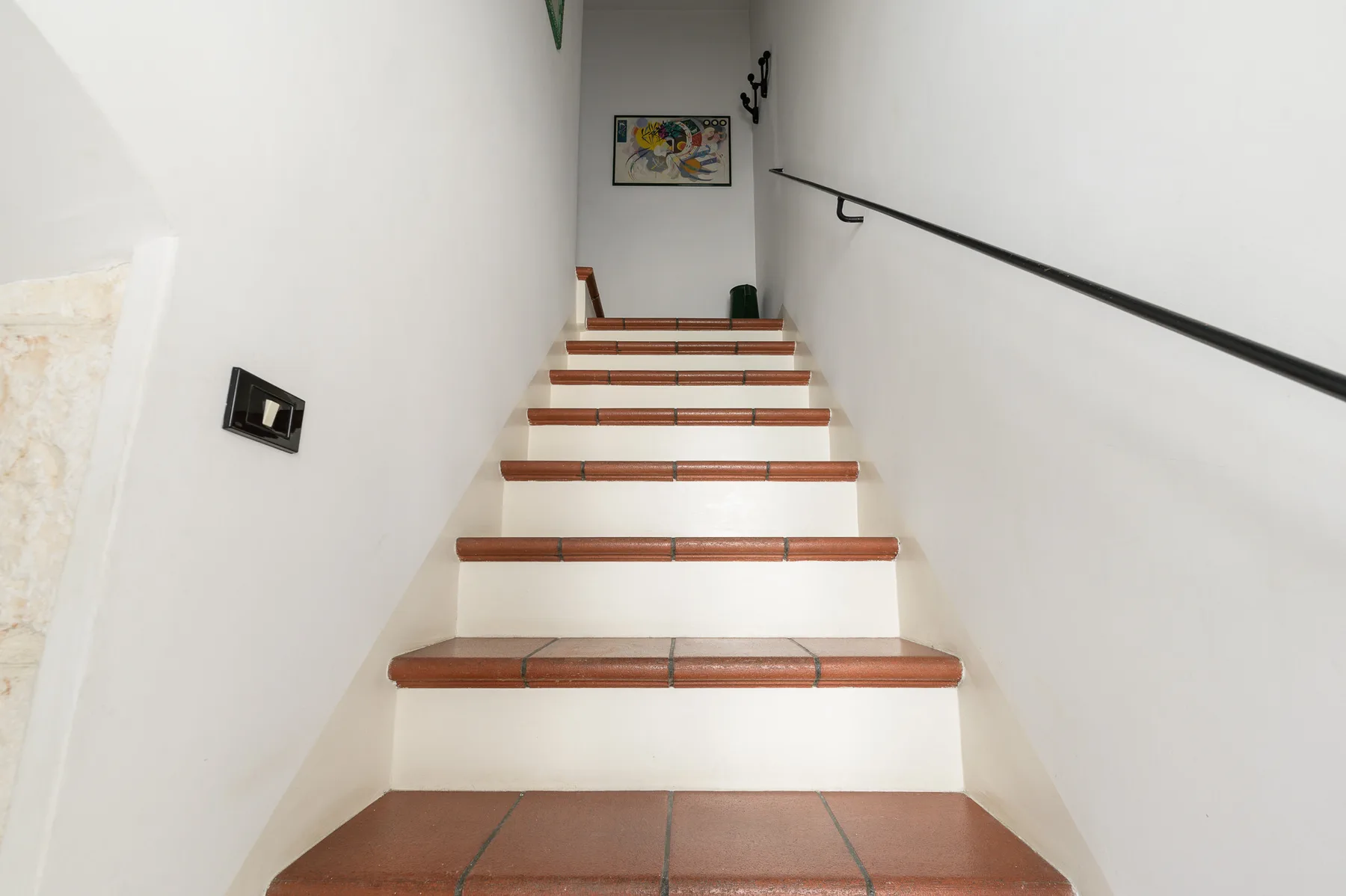 Una escalera de baldosas sube entre paredes blancas, con una barandilla oscura a un lado.