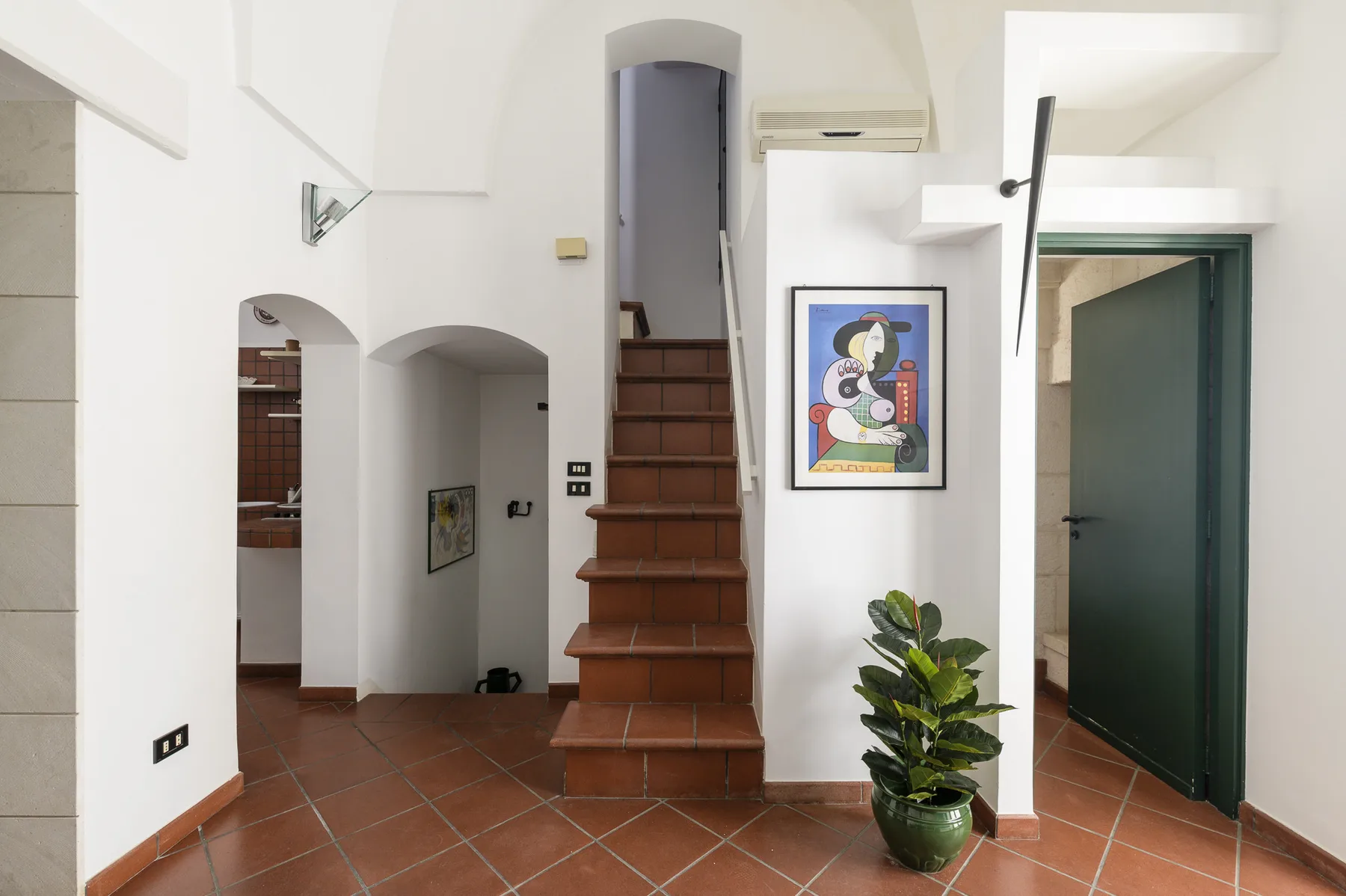 Un pasillo interior luminoso con una escalera, una obra enmarcada y una planta en maceta.