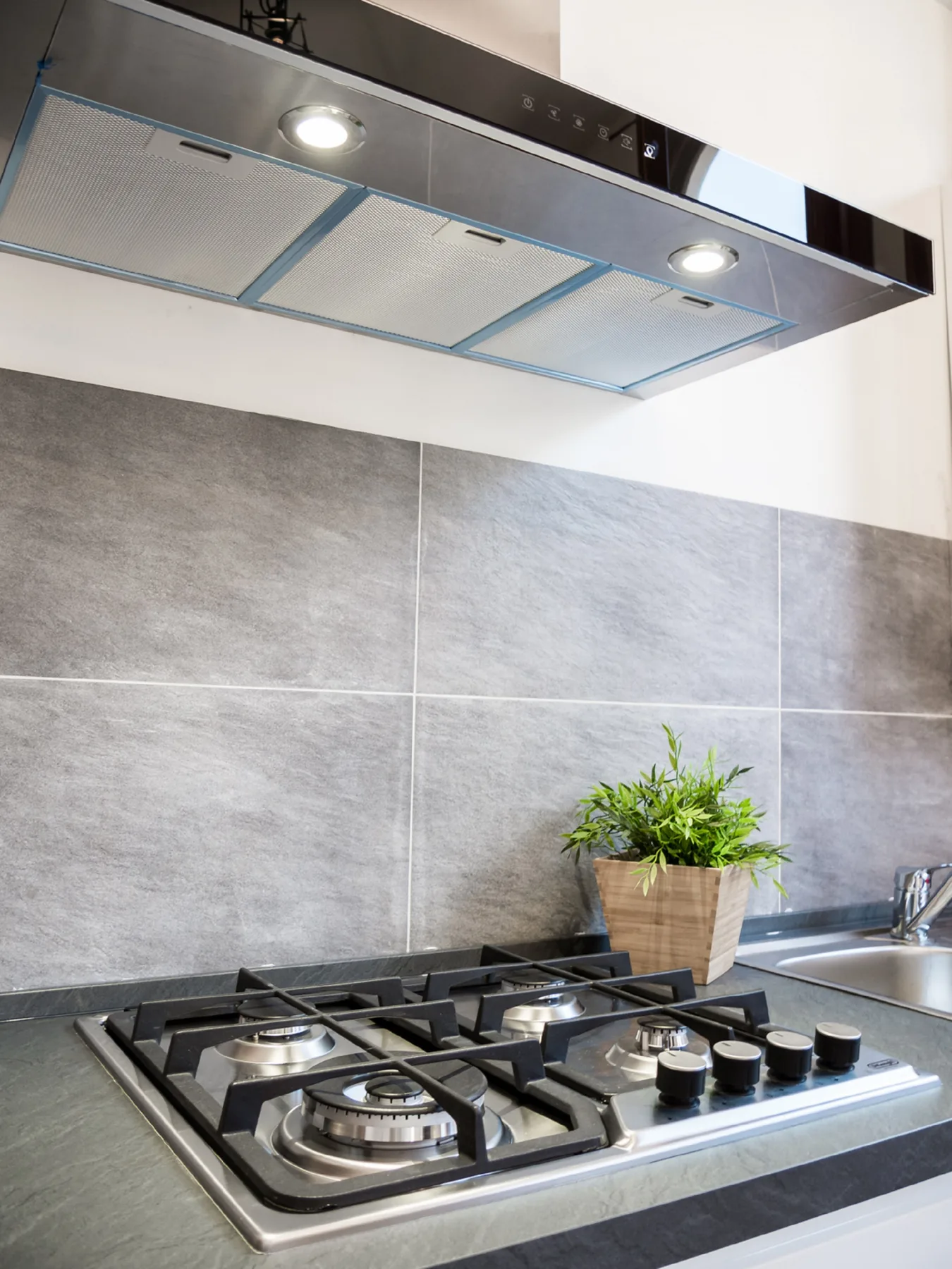 A kitchen cooktop sits beneath a stainless range hood against a tiled backsplash.