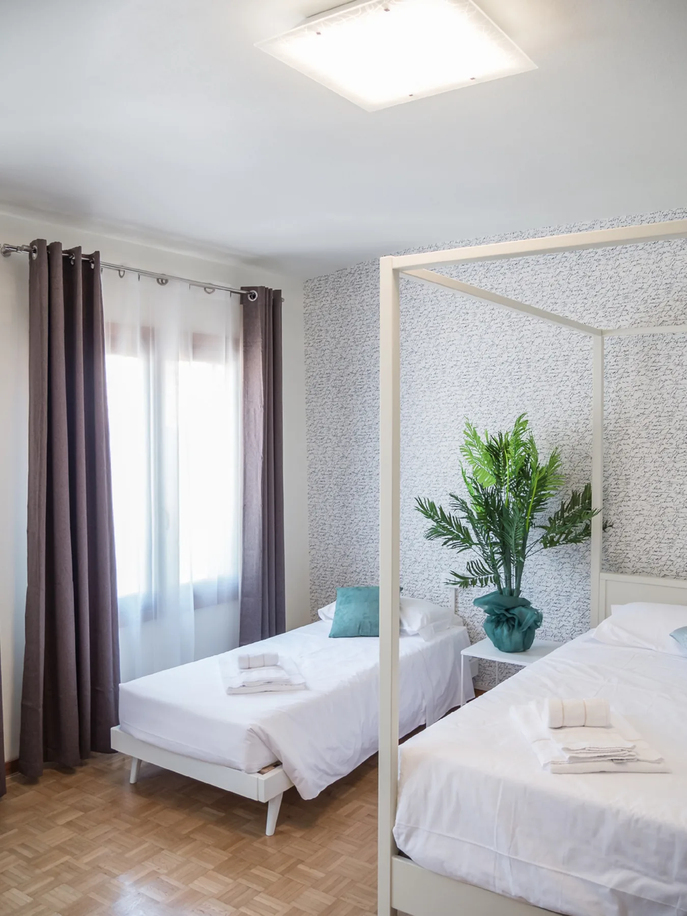 A bright bedroom with two beds, curtained windows, and a potted plant.
