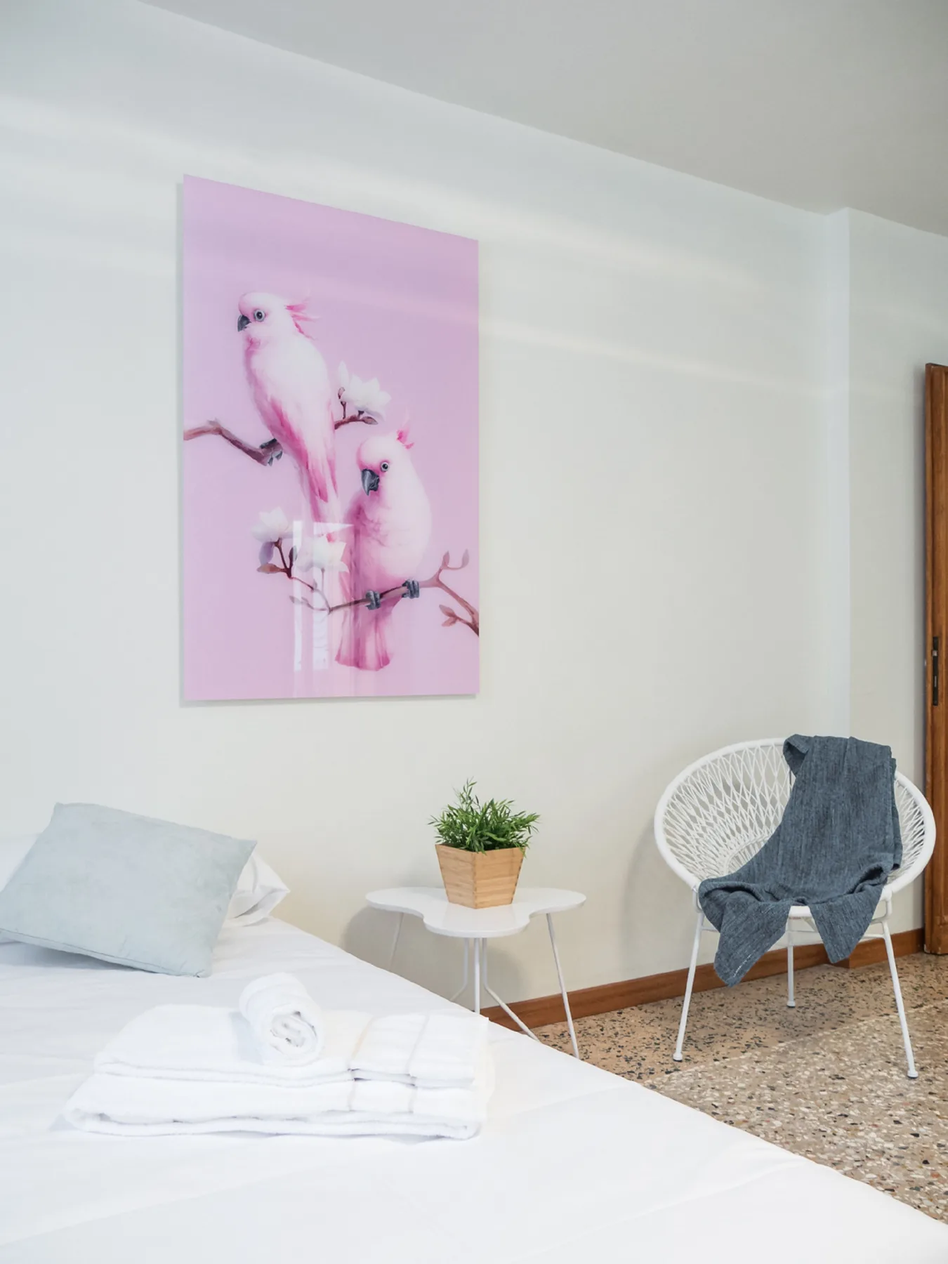 A bright bedroom with white bedding, a small side table, and pink bird artwork.