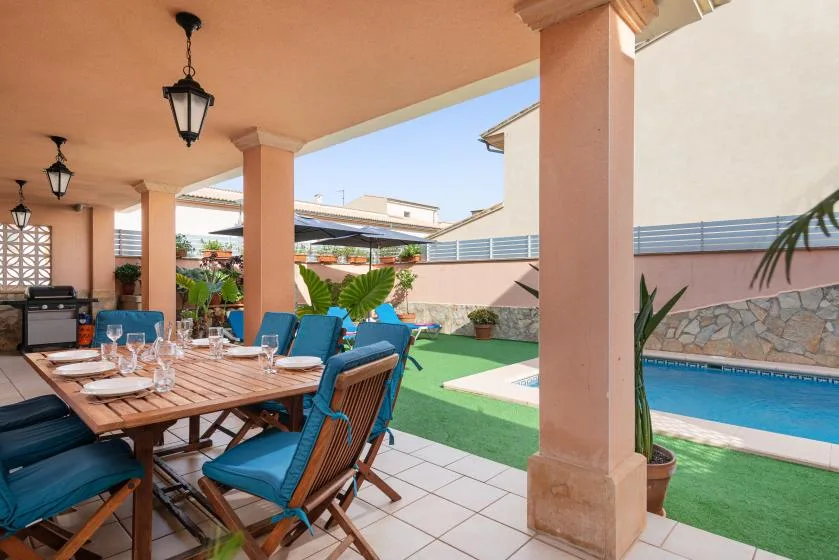 A covered patio has a dining table beside a swimming pool and green lawn.