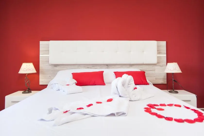 A bedroom with white bedding, red pillows, and rose petals arranged on the bed.
