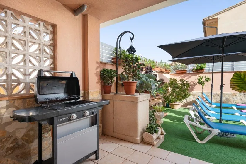 An outdoor patio with a barbecue grill, potted plants, and lounge chairs under a large umbrella.