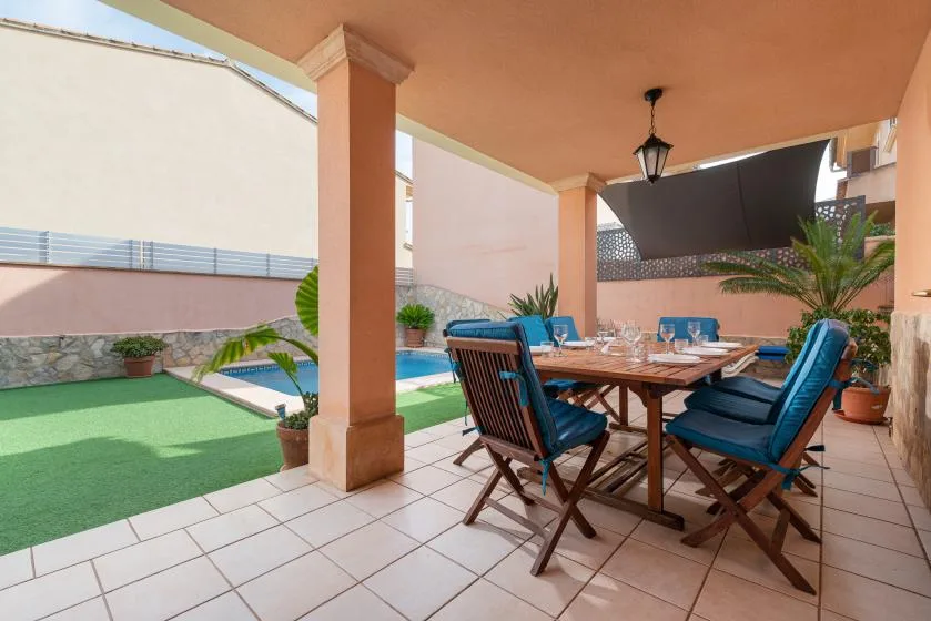 A covered patio dining area with a wooden table and blue seating beside a pool.