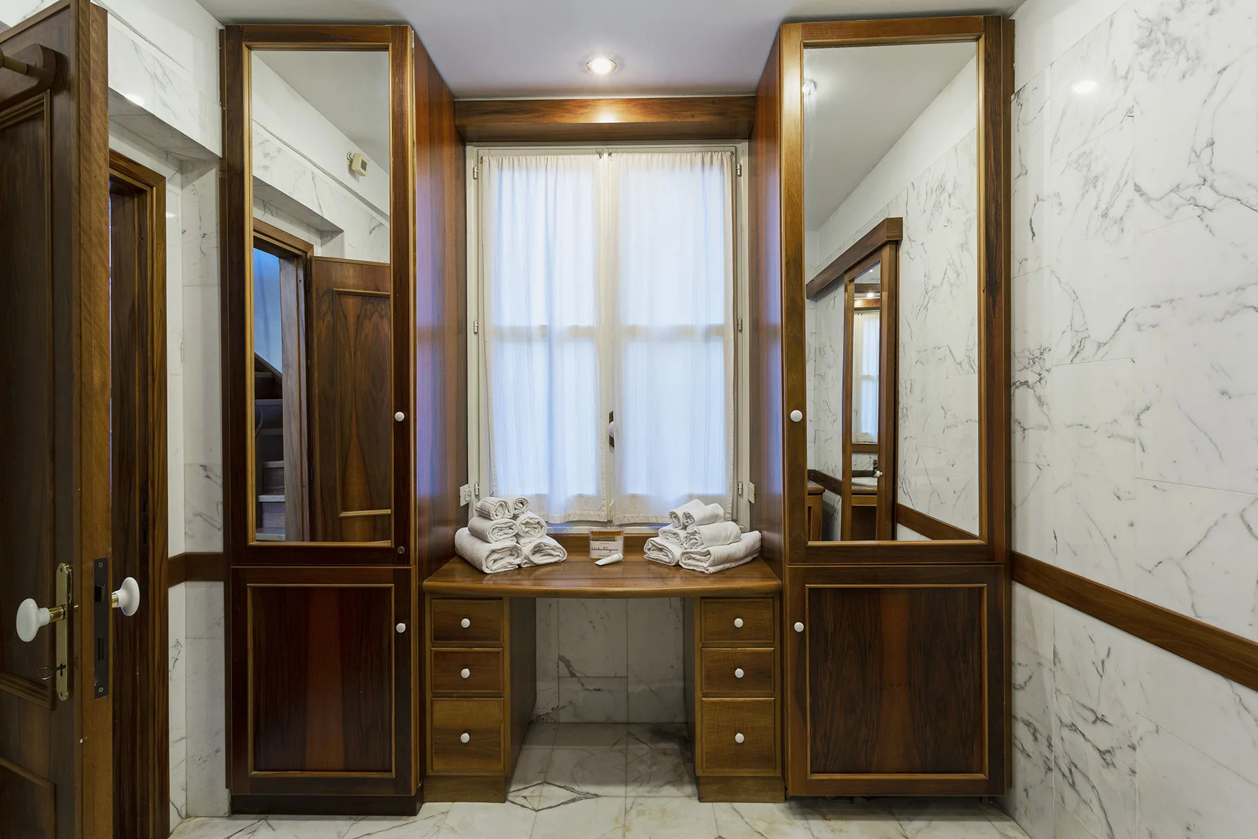 A dressing area with built-in wooden cabinetry, a curtained window, and folded towels.