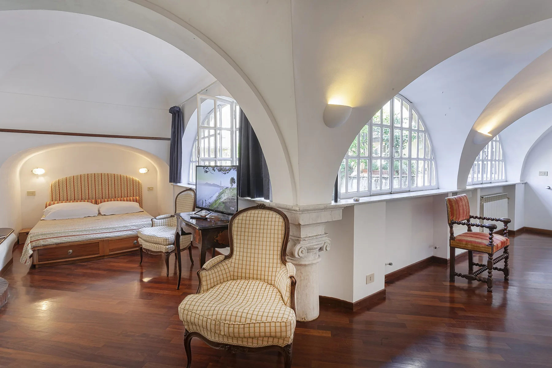 A bedroom and sitting area share a room with arched ceilings and large windows.