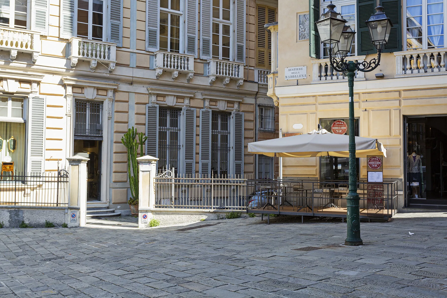 A paved courtyard fronts a pale building with shuttered windows and an outdoor canopy.