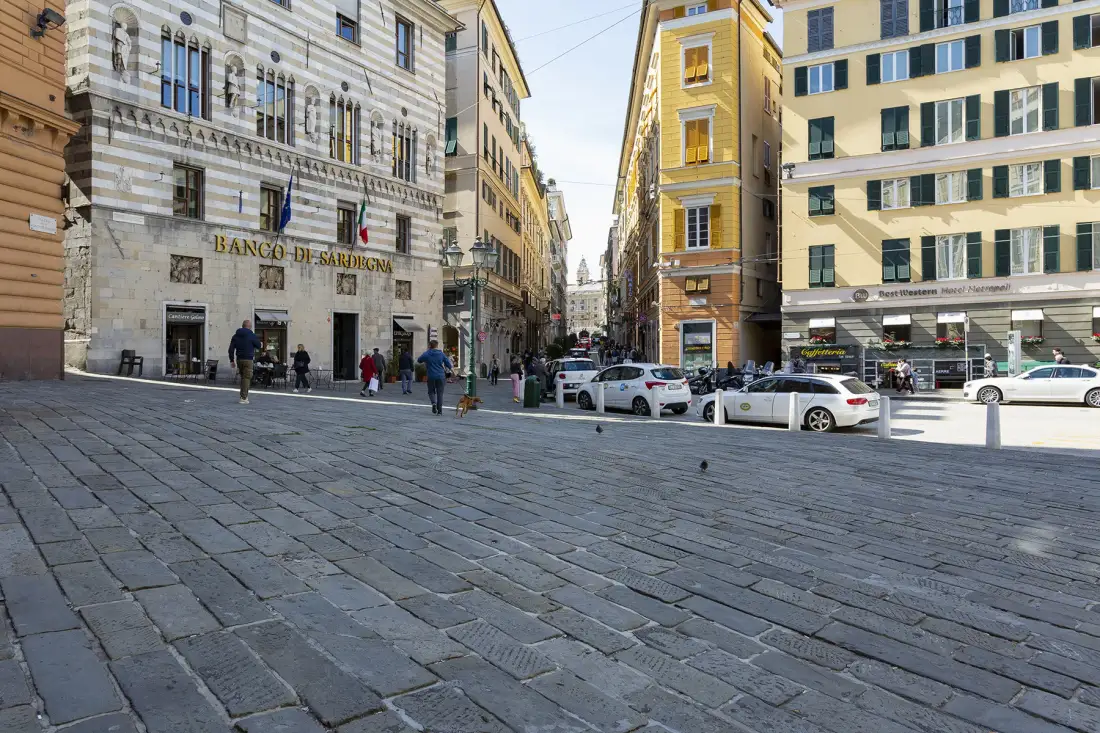 A paved city square bordered by multistory buildings, with pedestrians and parked cars.