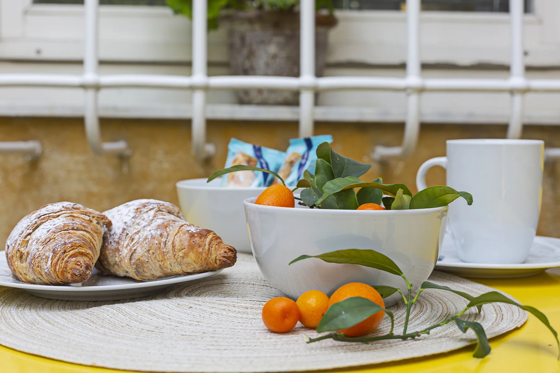 A breakfast table displays pastries, citrus fruit, and a white cup beside a bowl of greenery.