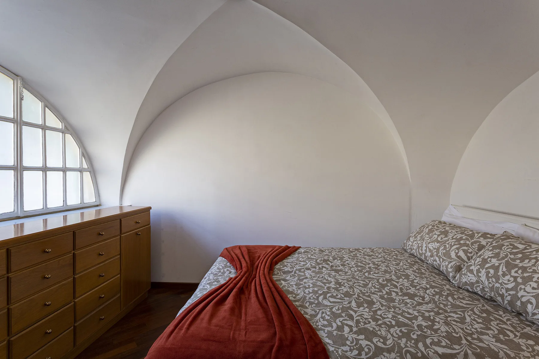 A bedroom with a patterned bed, red throw, and wooden dresser beneath an arched window.