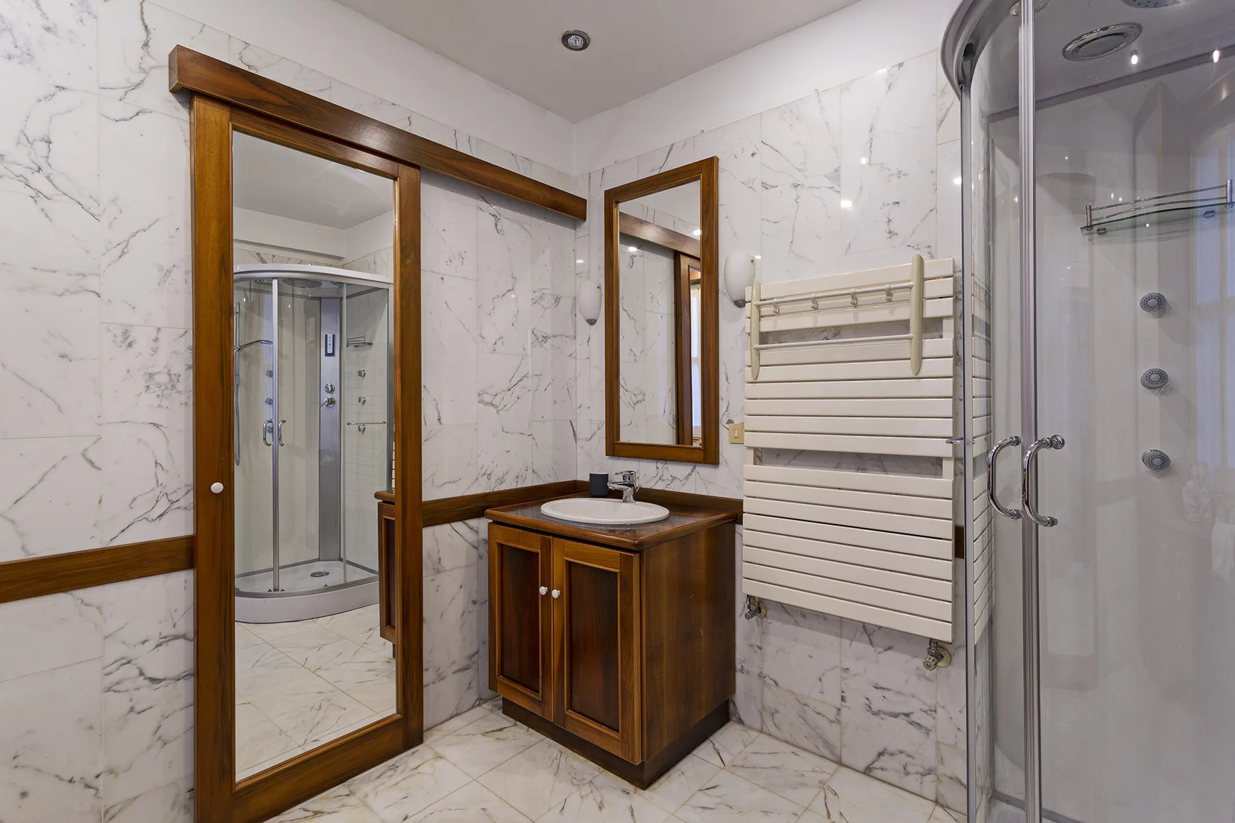 A tiled bathroom with a vanity, wall-mounted towel warmer, and glass shower.