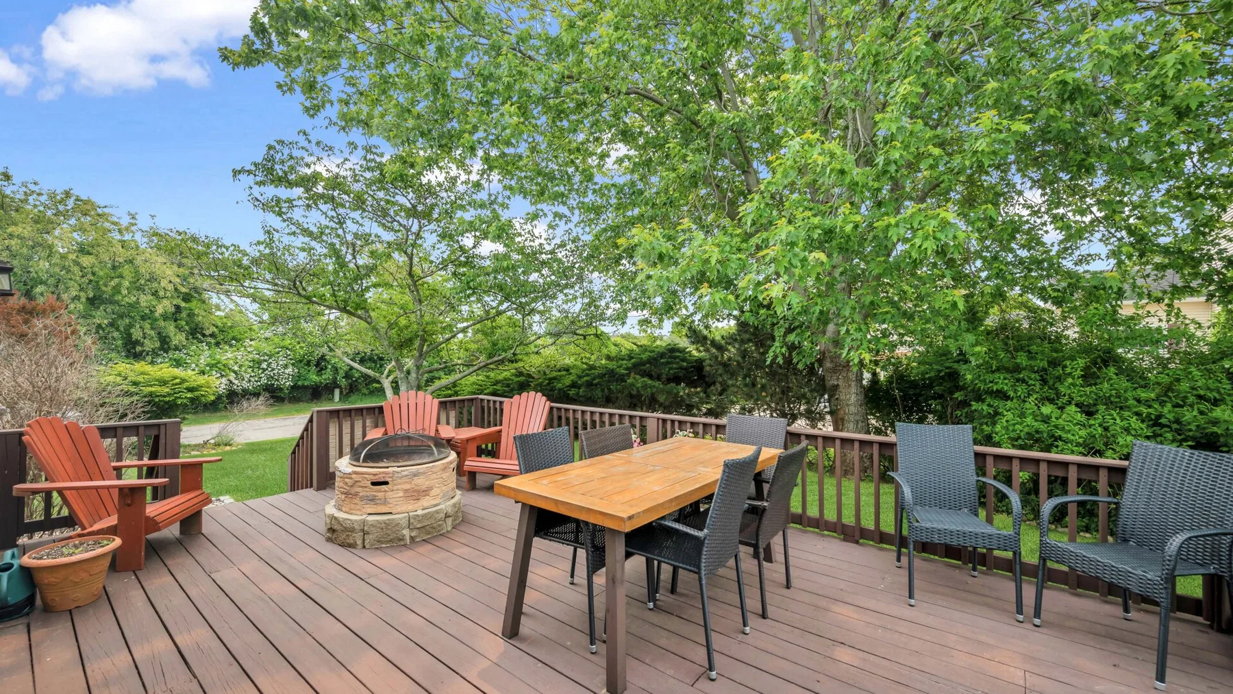 An outdoor deck with a wooden dining table and seating beneath leafy trees.
