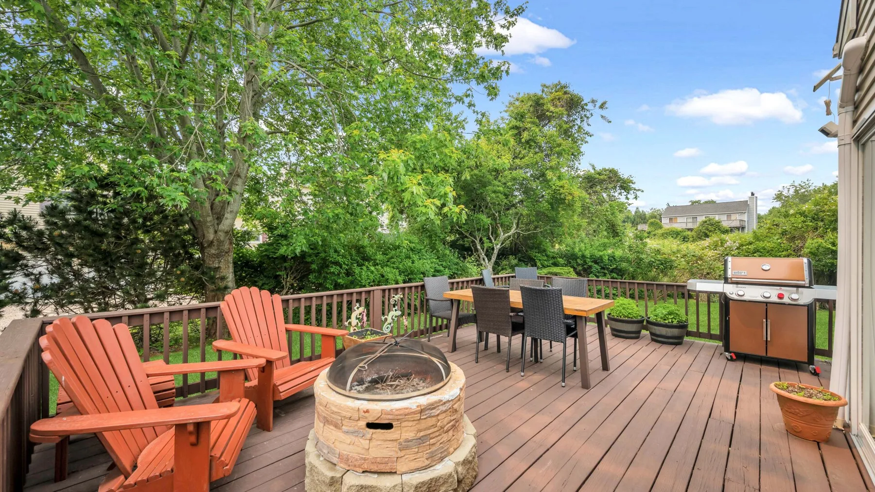 Una terraza de madera al aire libre tiene asientos para comer, un brasero y una parrilla, rodeados de árboles.