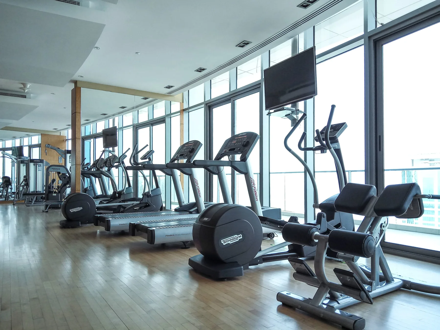 Un gimnasio con máquinas de cardio junto a grandes ventanas.