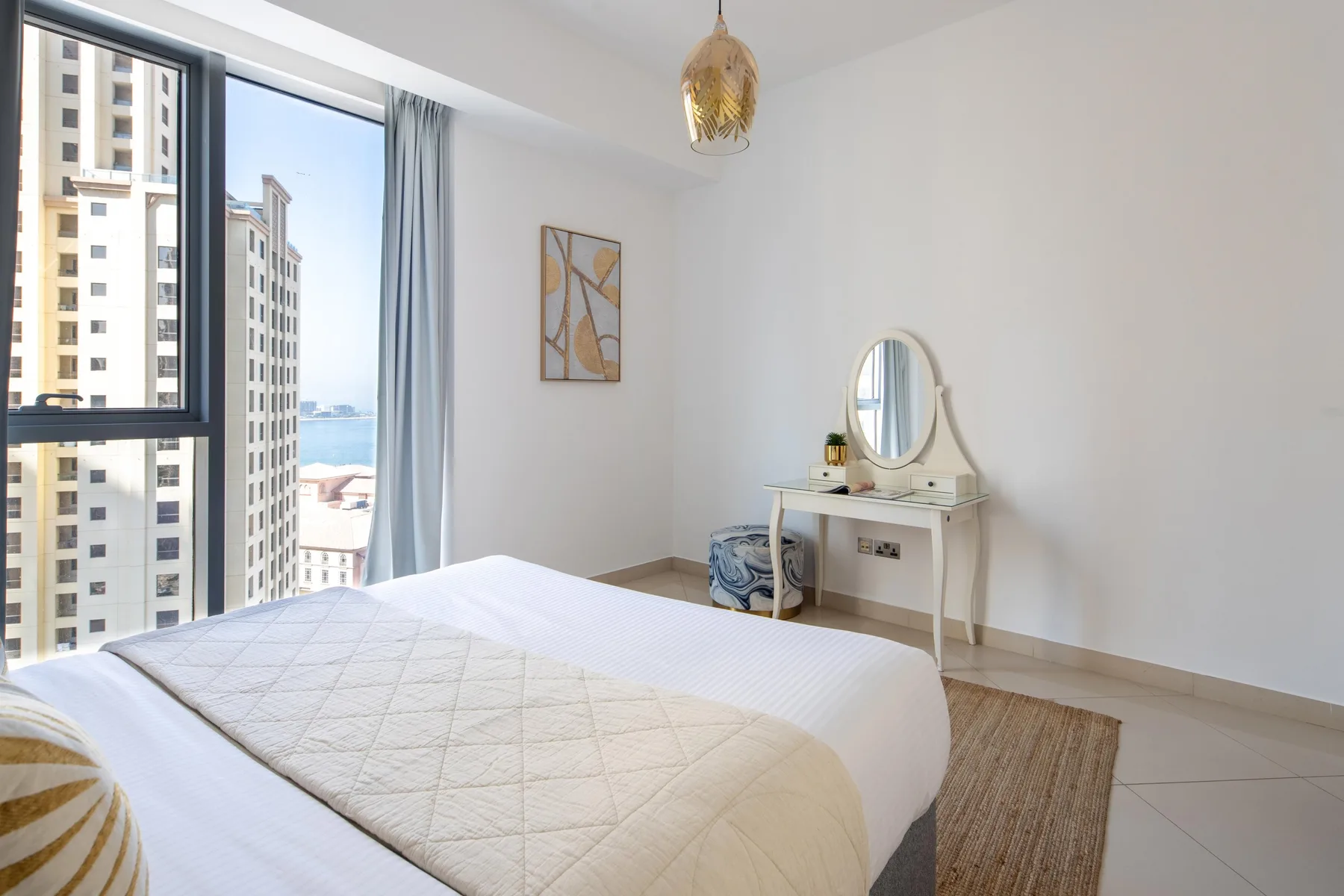 Un dormitorio luminoso con una cama, un tocador y ventanas grandes.