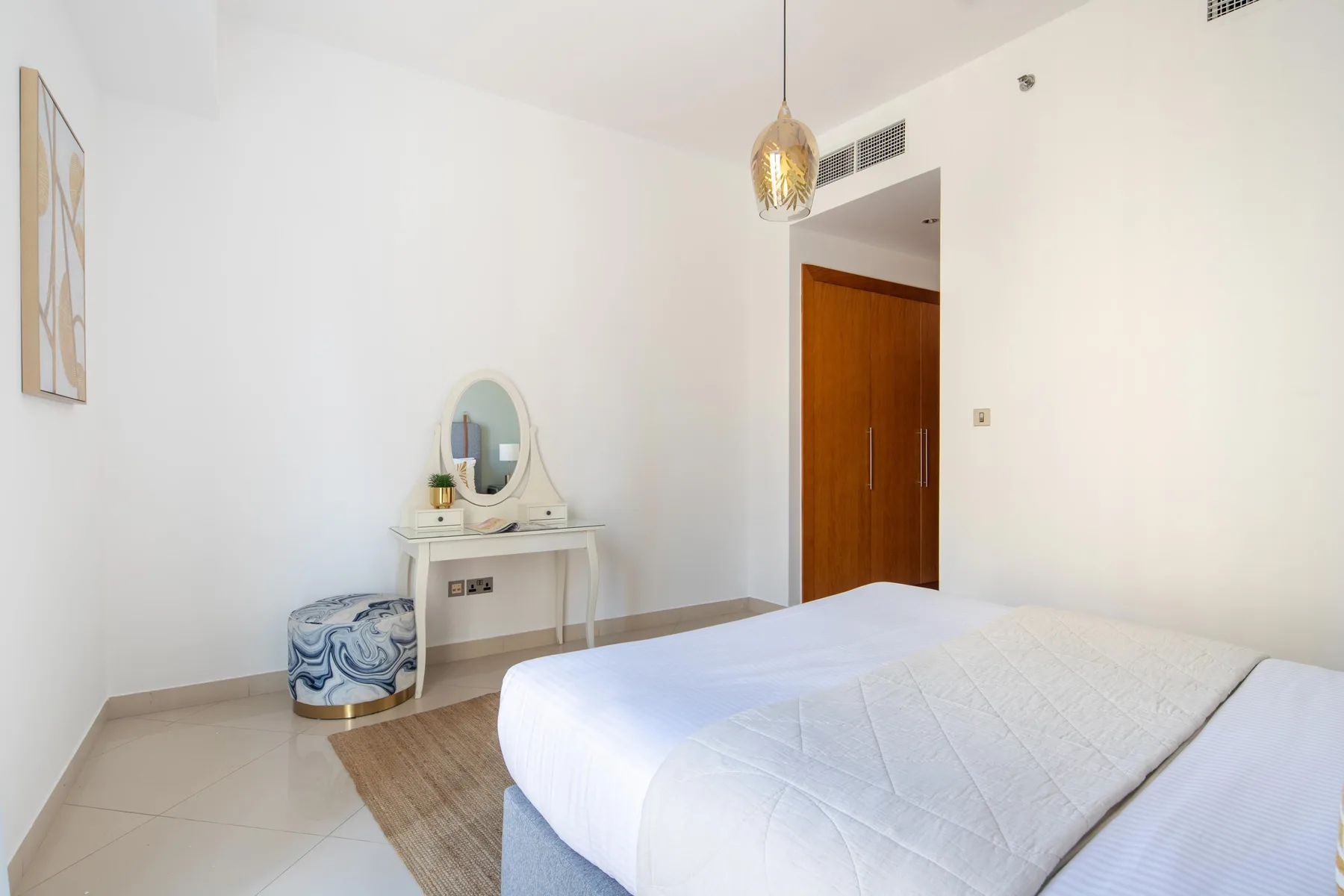 Un dormitorio luminoso con una cama, un pequeño tocador y una puerta de madera.