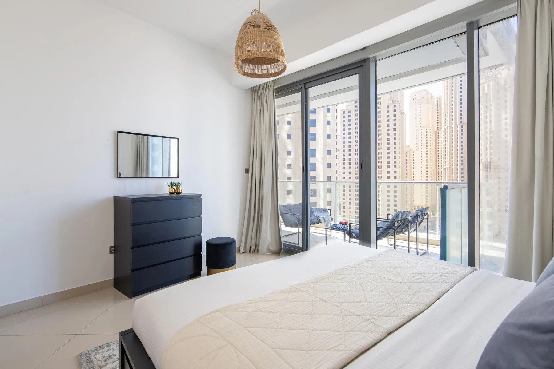 Un dormitorio luminoso con una cama, una cómoda oscura y puertas de vidrio que dan a un balcón.