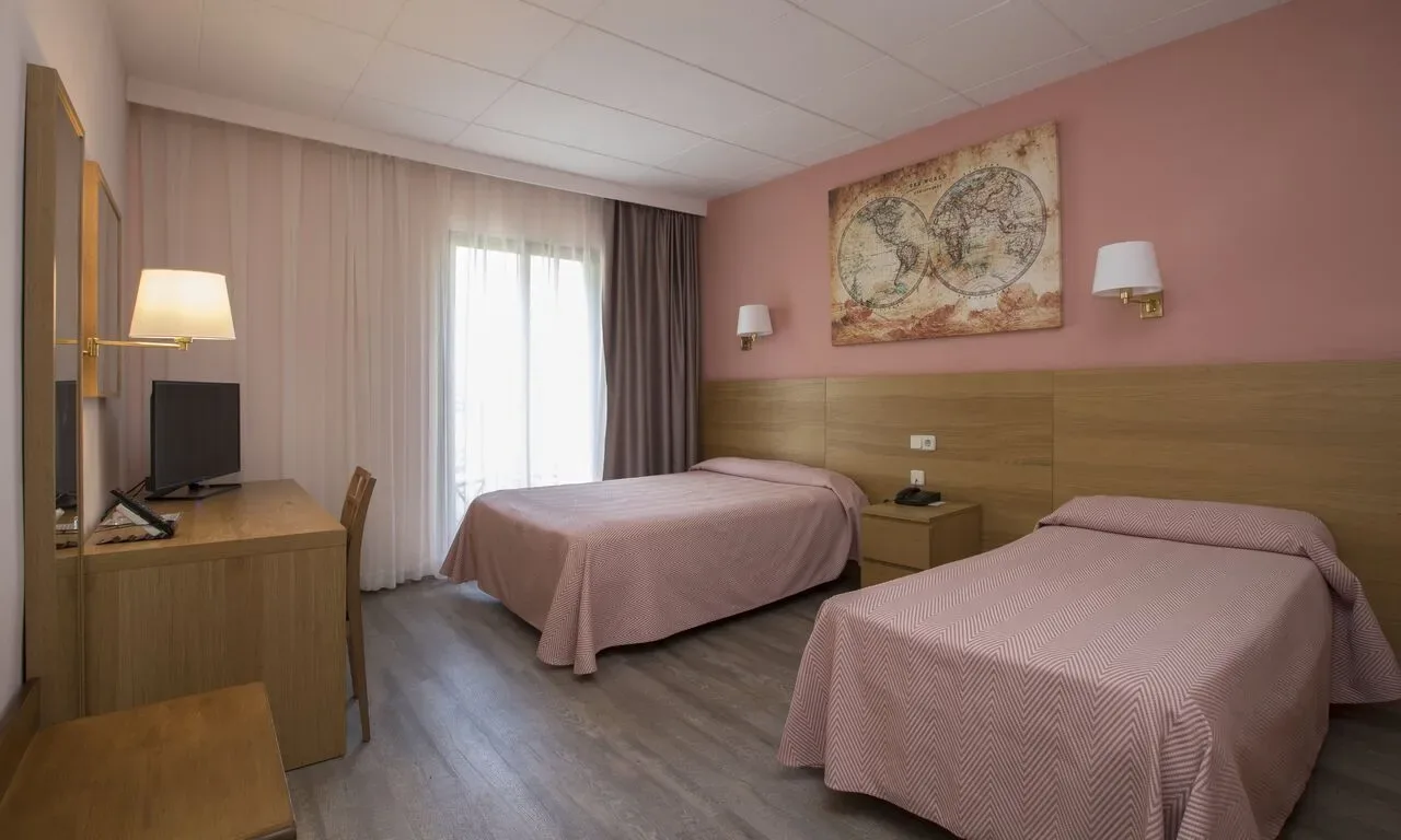 Habitación de hotel con dos camas individuales, un escritorio y obras de arte en una pared rosa.