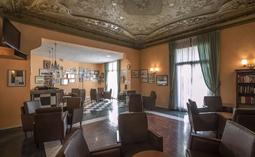 Habitación de hotel con sillas marrones, suelo a cuadros, techo ornamentado y fotos enmarcadas.