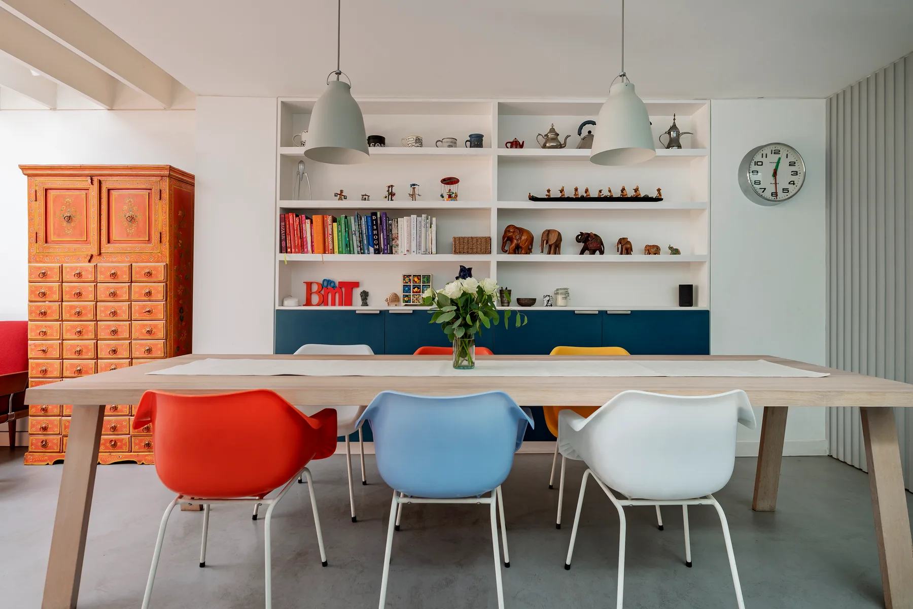 Un comedor con una mesa larga, asientos coloridos y estantes en la pared del fondo.