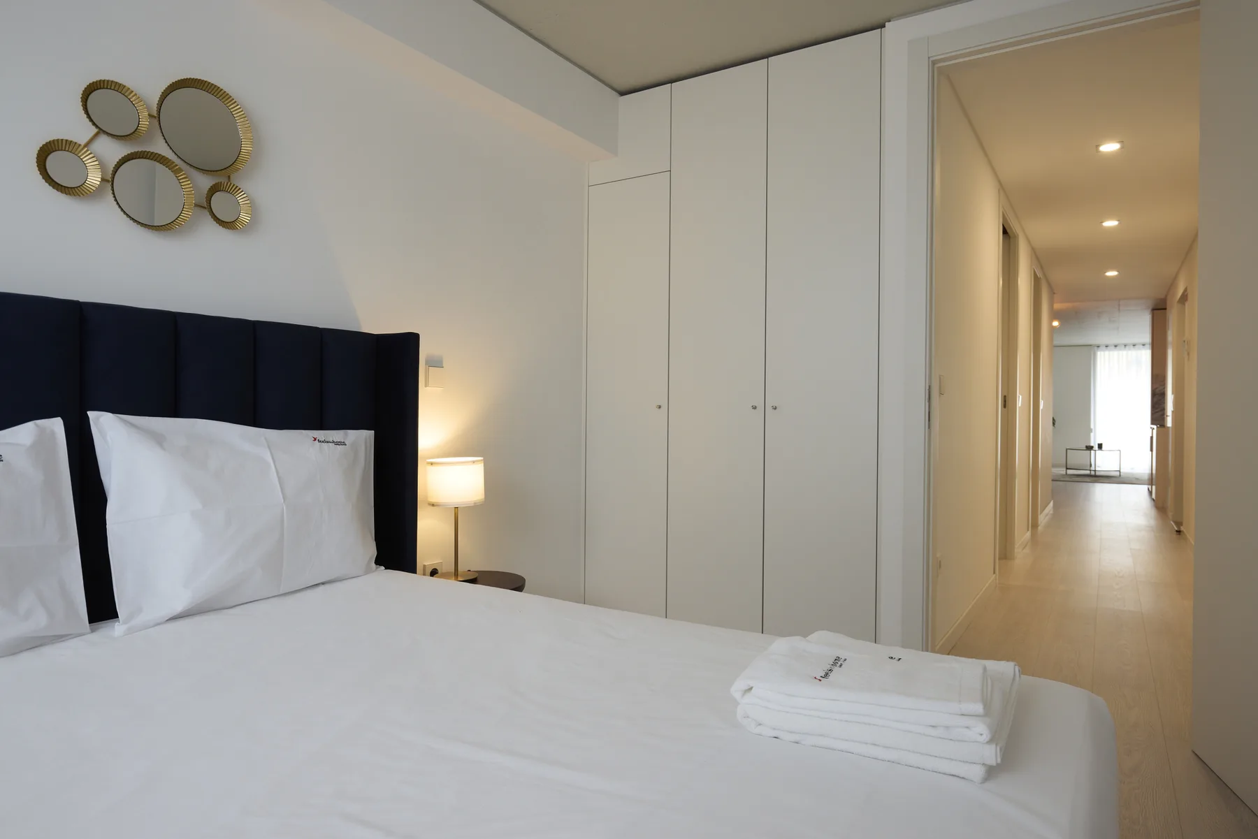 A bedroom with a neatly made bed and a hallway leading toward a bright window.