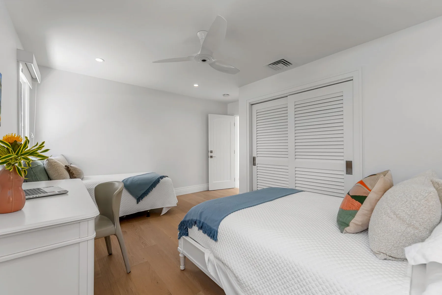 Un dormitorio luminoso con dos camas, puertas de armario con persianas y un ventilador de techo.