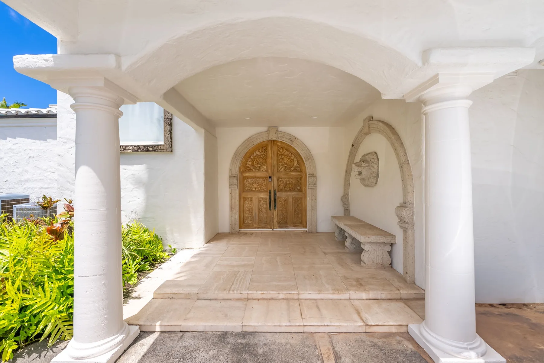 Una entrada blanca con arco, puertas dobles de madera ornamentadas y un banco de piedra.
