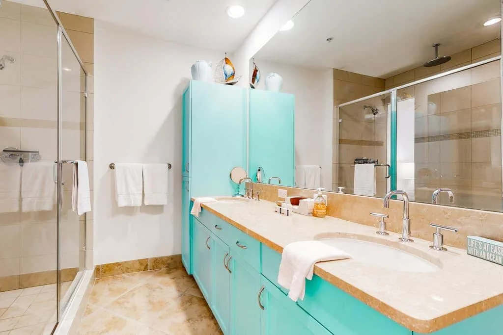 A bright bathroom has turquoise cabinetry and a long counter with sinks.