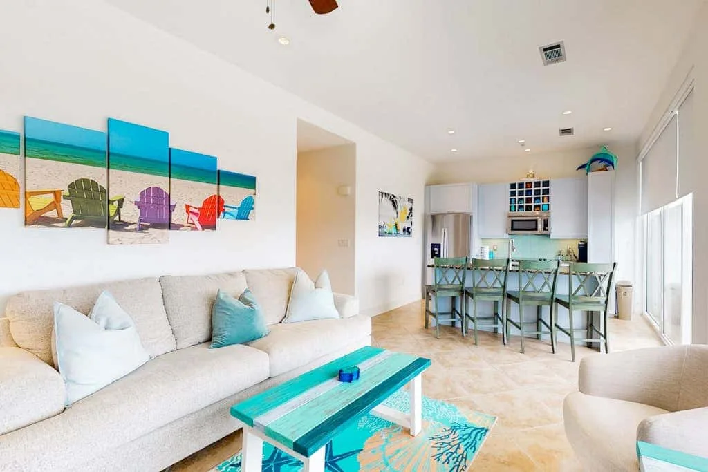 A bright open living area with seating, a turquoise coffee table, and a kitchen counter.