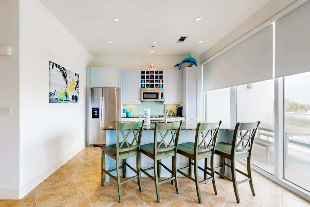 A bright dining area with a long table and large windows beside a compact kitchen.