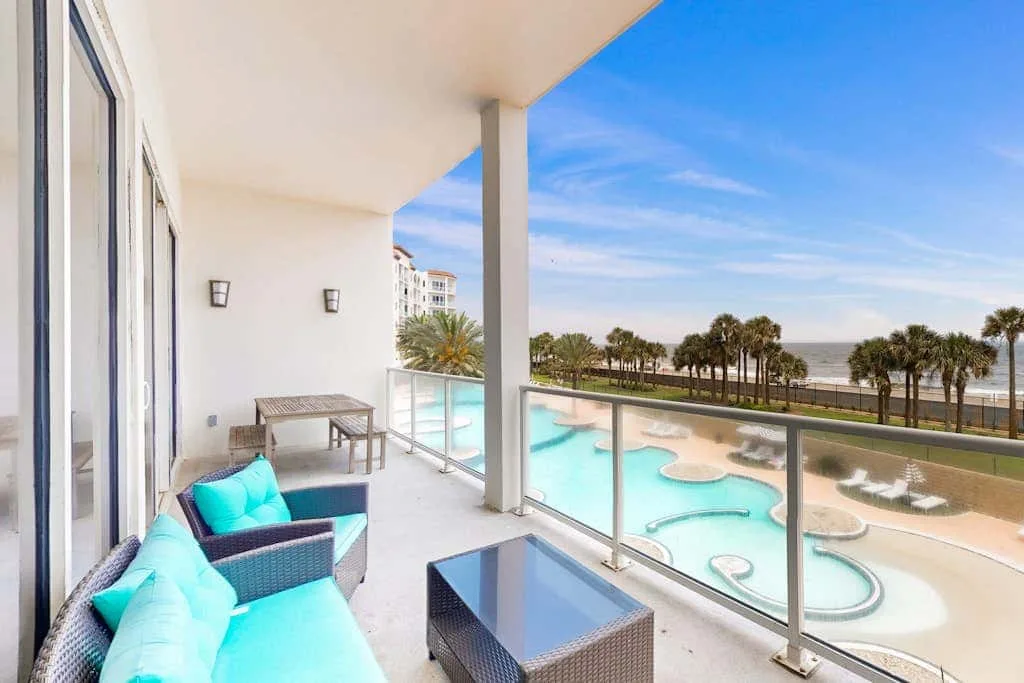 A balcony seating area overlooks a turquoise pool and palm trees.