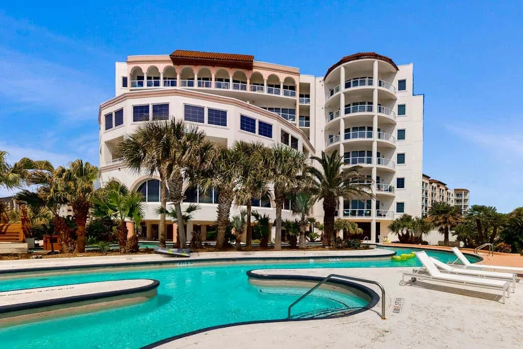 A curved outdoor pool sits in front of a multi-story building lined with palm trees.
