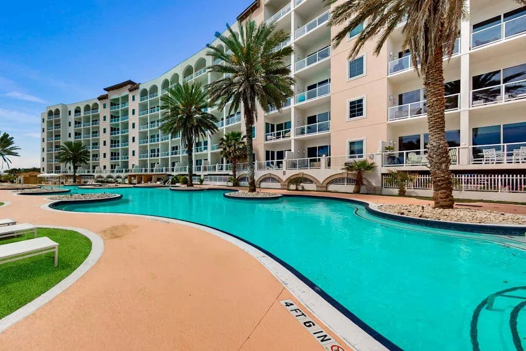A curved outdoor pool sits beside a multi-story building and palm trees.