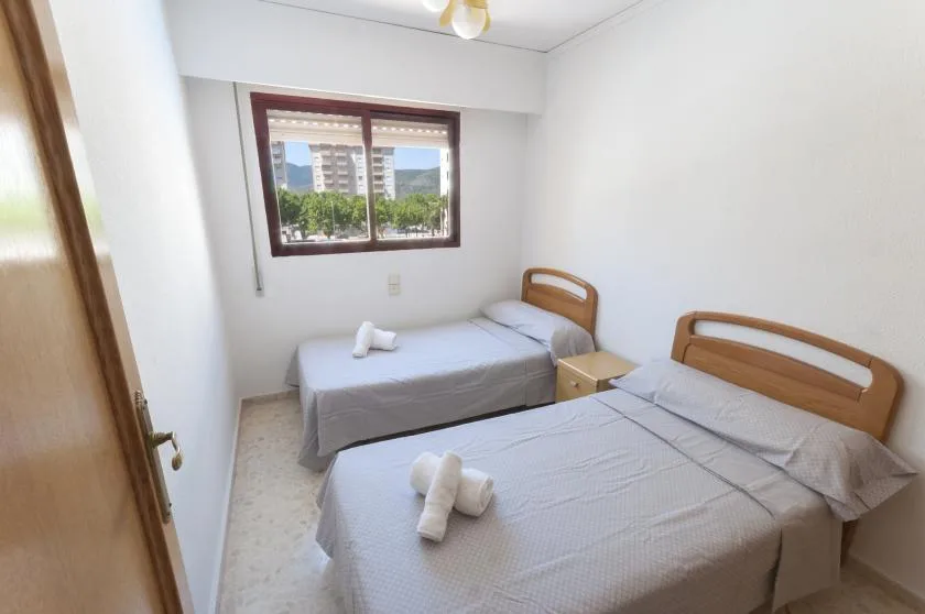 A bright bedroom with two beds and a window overlooking nearby buildings.