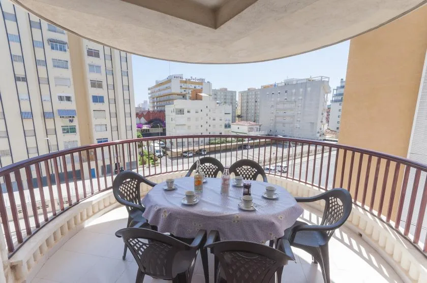 A covered balcony has a round table with seating and views of nearby buildings.