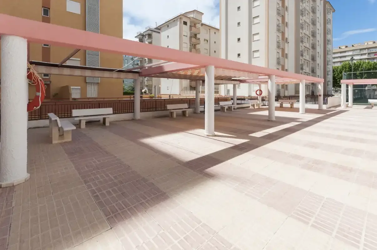 A paved outdoor terrace with benches beneath a pink canopy, surrounded by apartment buildings.