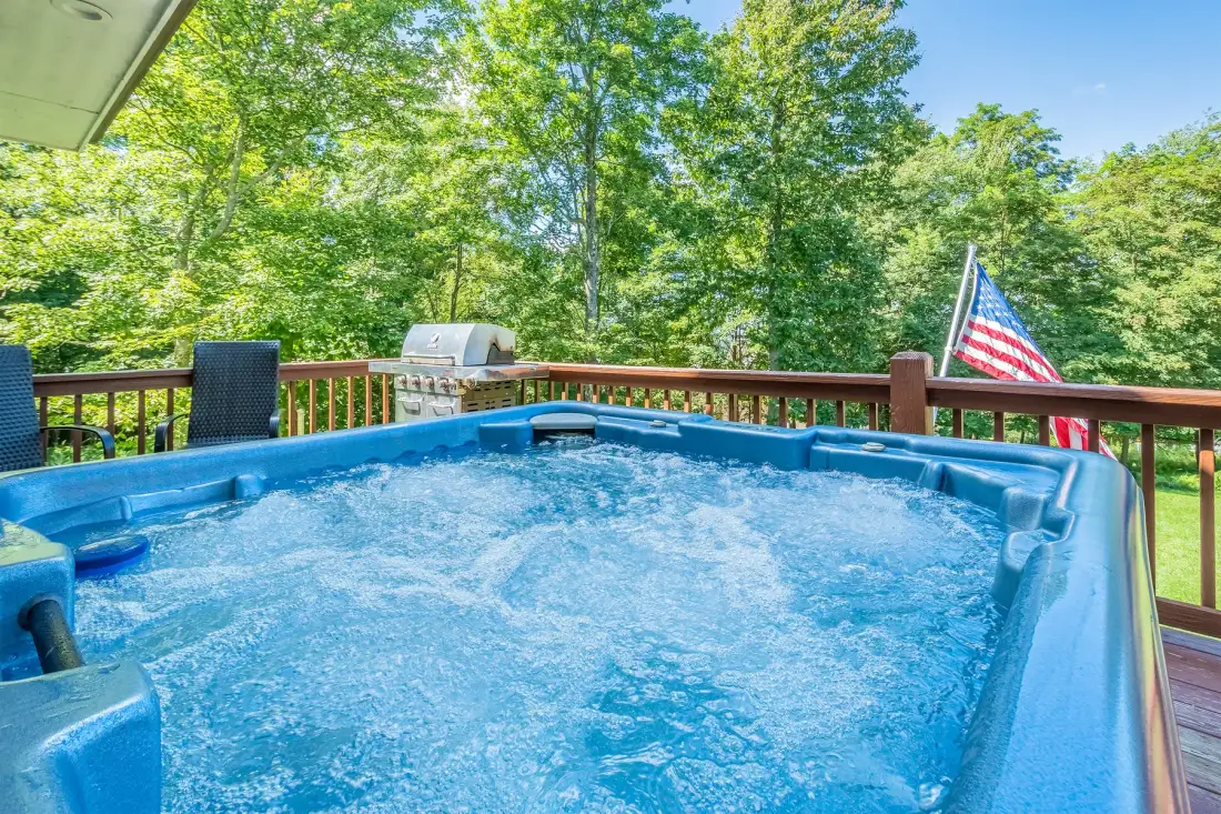 An outdoor hot tub bubbles on a wooden deck surrounded by leafy trees.