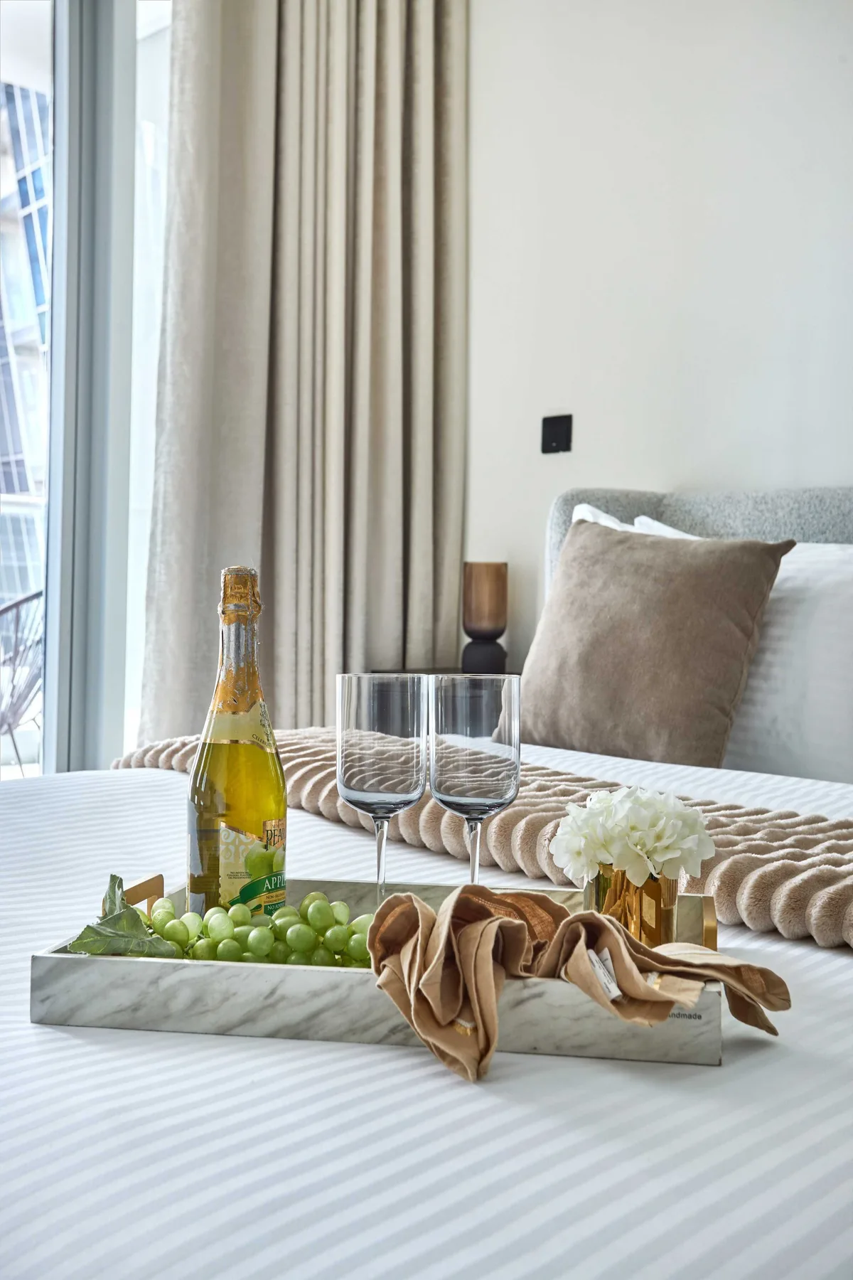 Una mesa de comedor tiene una bandeja con fruta y copas junto a una ventana con cortinas.