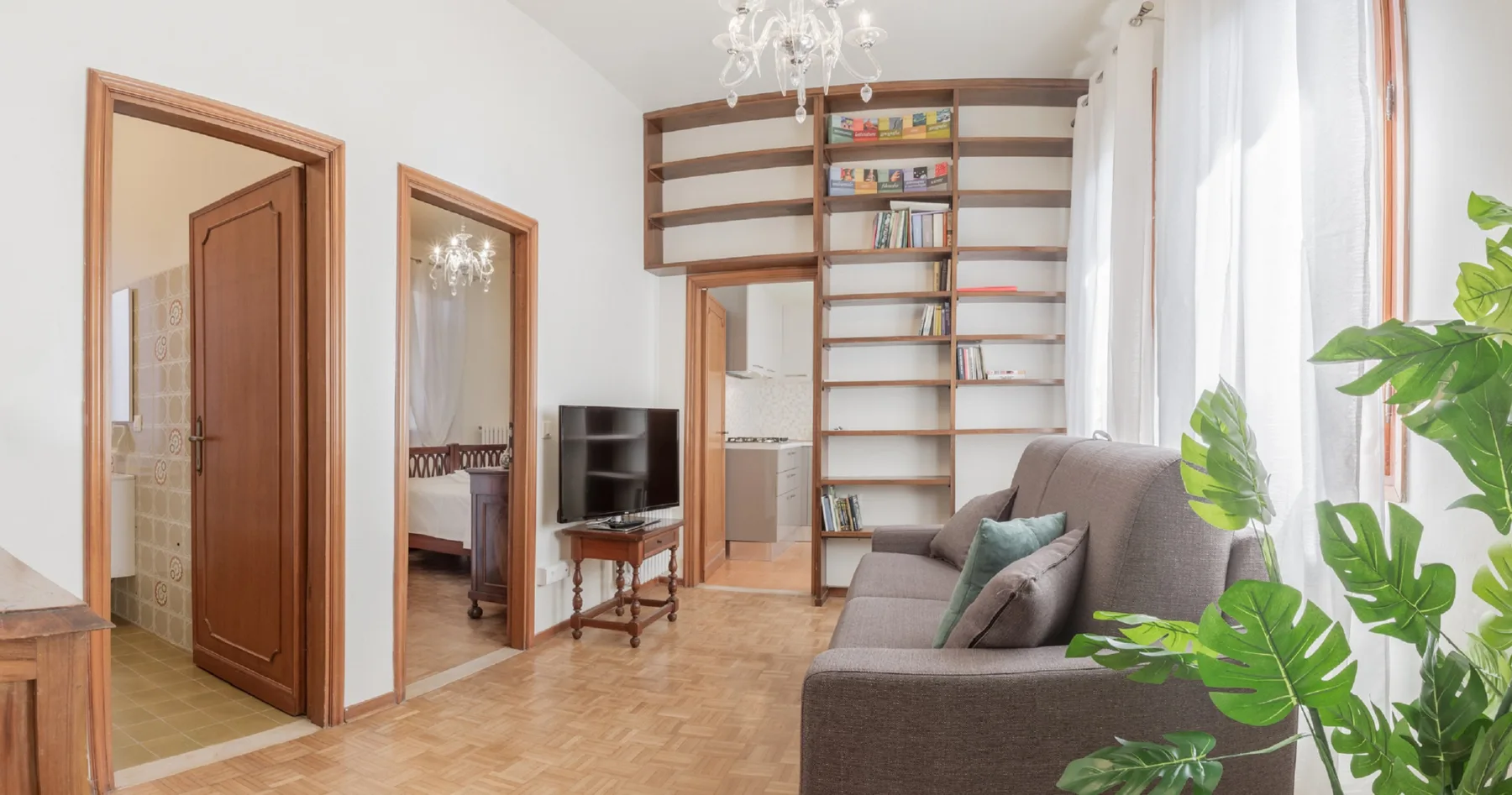 Una sala luminosa con sofá gris, televisor y estanterías en la pared.