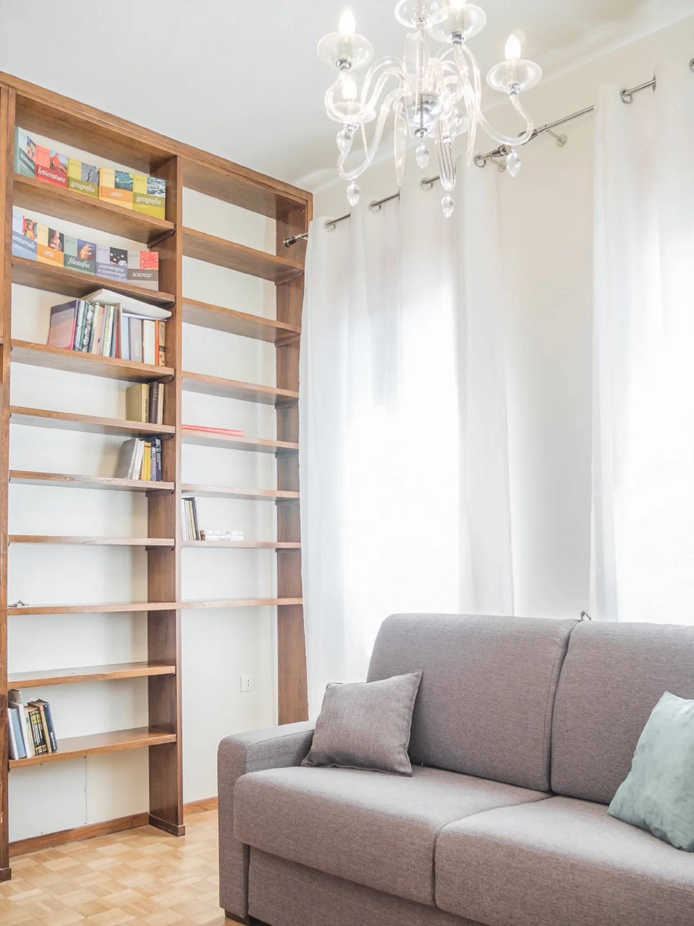 Una sala luminosa con un sofá gris, estanterías de madera y cortinas blancas.