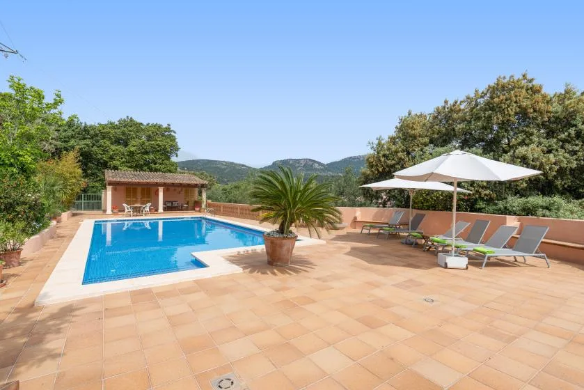 Una zona de piscina al aire libre tiene una terraza pavimentada, asientos reclinables y una sombrilla.