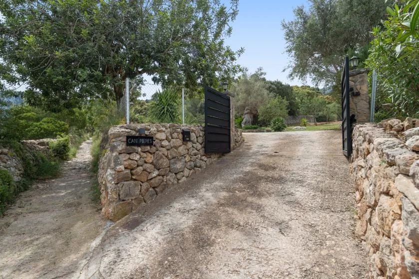 Un camino de grava pasa por una puerta abierta entre muros bajos de piedra y árboles.