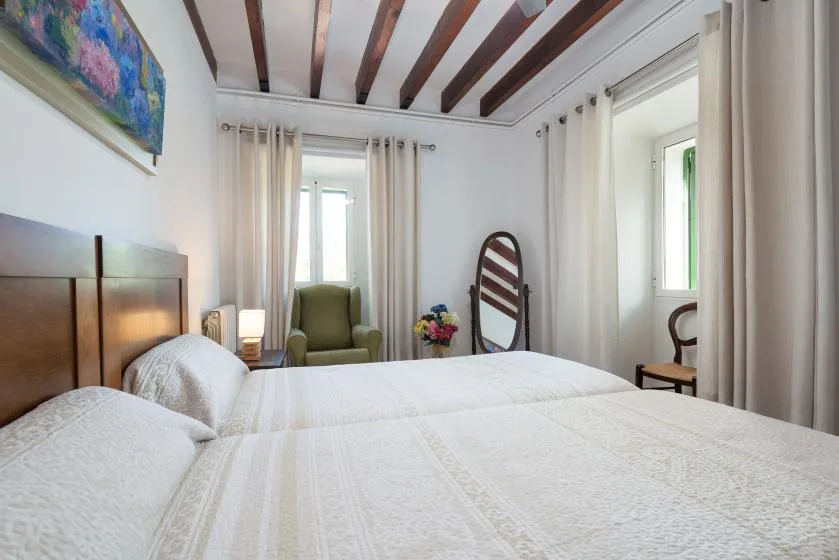 Un dormitorio luminoso con ropa de cama blanca, ventanas altas y vigas expuestas en el techo.
