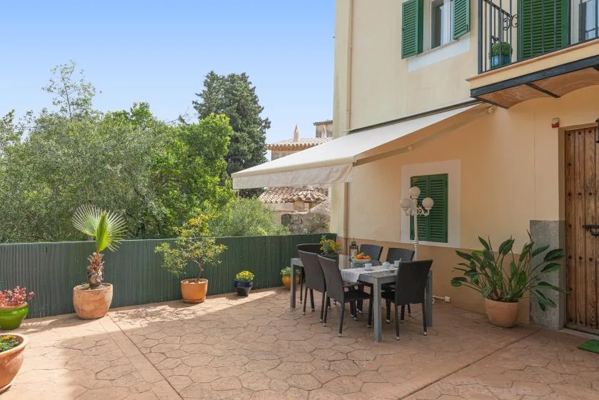 Un patio exterior pavimentado con mesa de comedor, asientos y plantas en macetas junto a una casa.