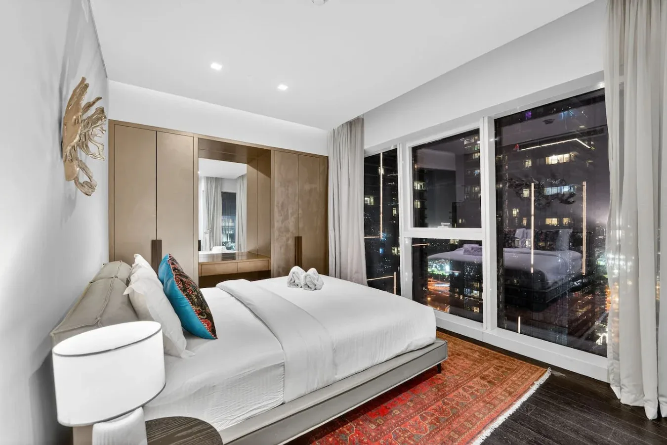 A bright bedroom with a neatly made bed beside large windows overlooking city lights.